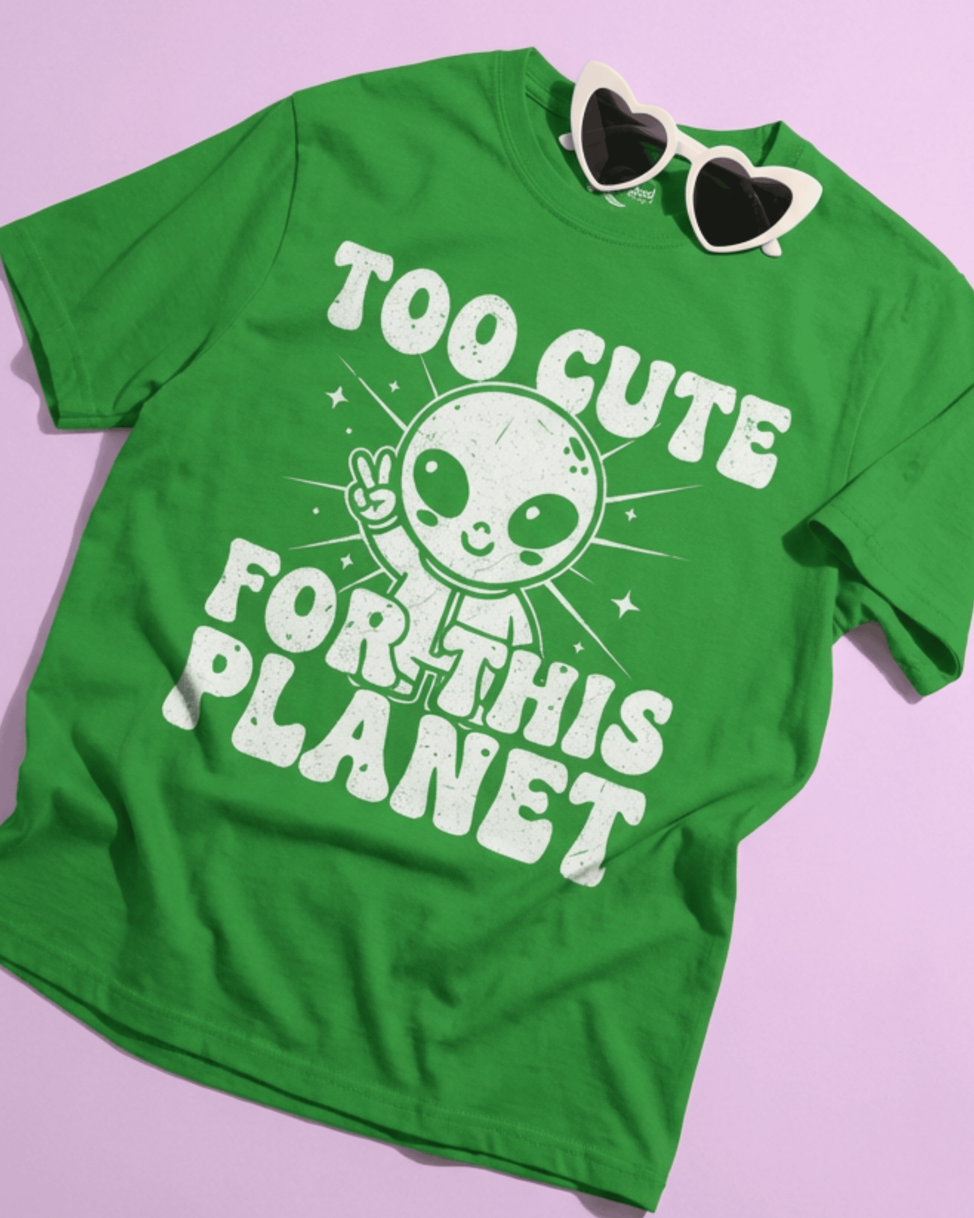A flat lay product shot of the green UFO Sci Fi Shirt on a solid pink background. This outer space lover t-shirt features the cute alien peace sign graphic and is styled with a pair of white Y2K-style heart sunglasses.