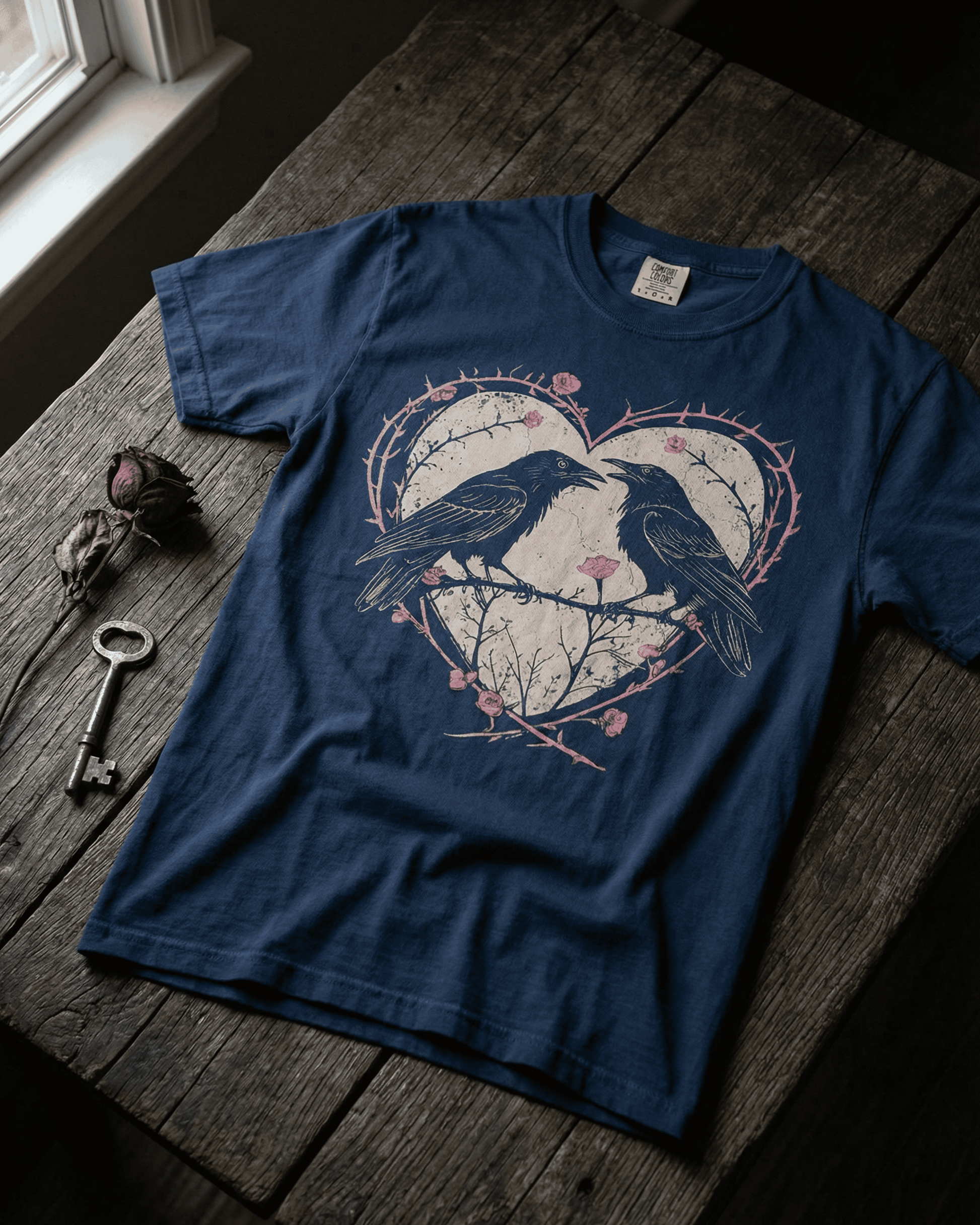 A navy Vintage Raven Comfort Colors Valentine T-Shirt laid on a rustic wooden surface. The grunge aesthetic graphic shows two crows within a heart of thorns and pink roses, embodying a Gothic Dark Romance theme.