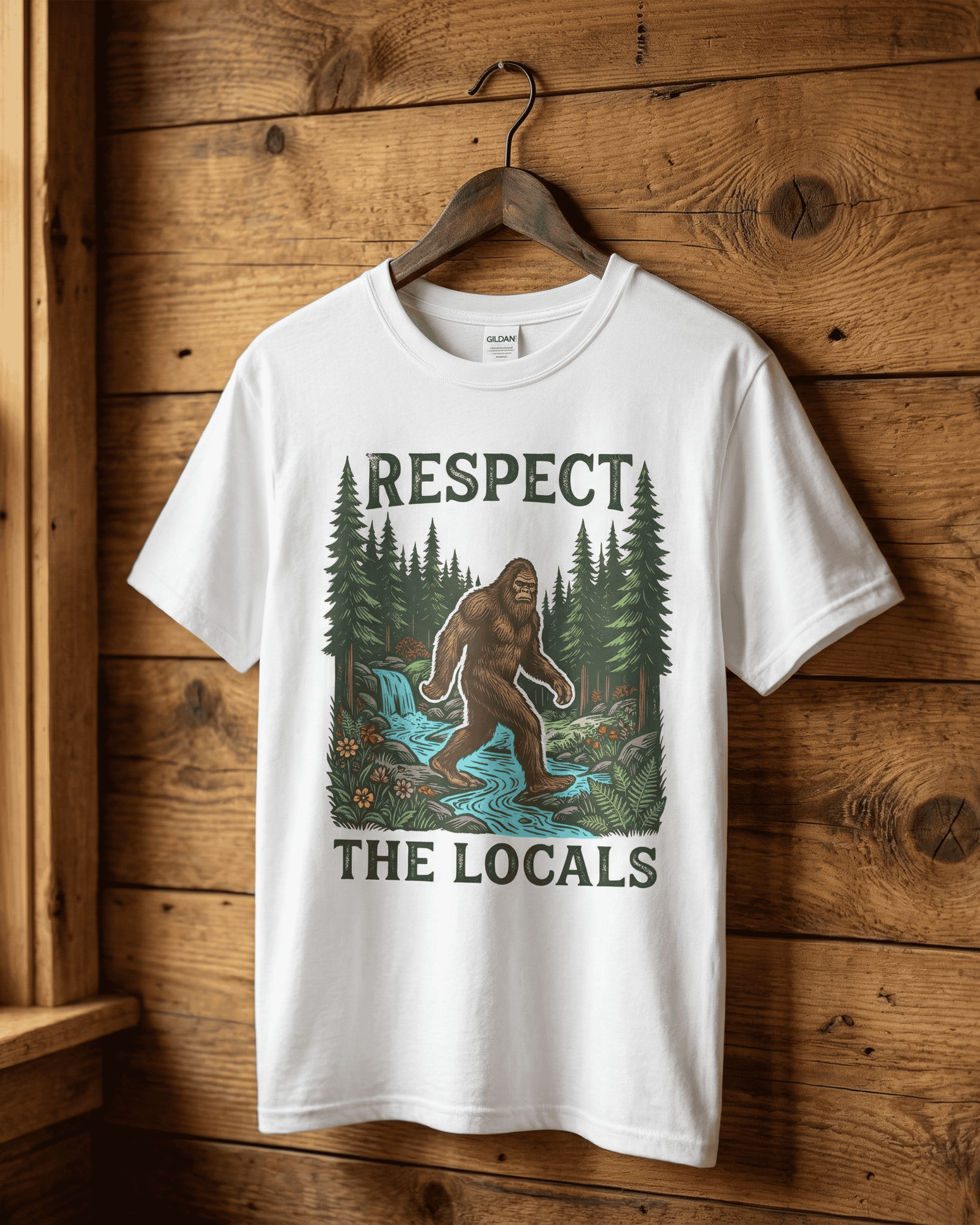 A crisp white 90s retro shirt featuring a Bigfoot 'Respect the Locals' design, hanging against a wood-paneled wall. The vintage-style graphic of Bigfoot in nature is ideal for camping trips and outdoor adventure lovers.