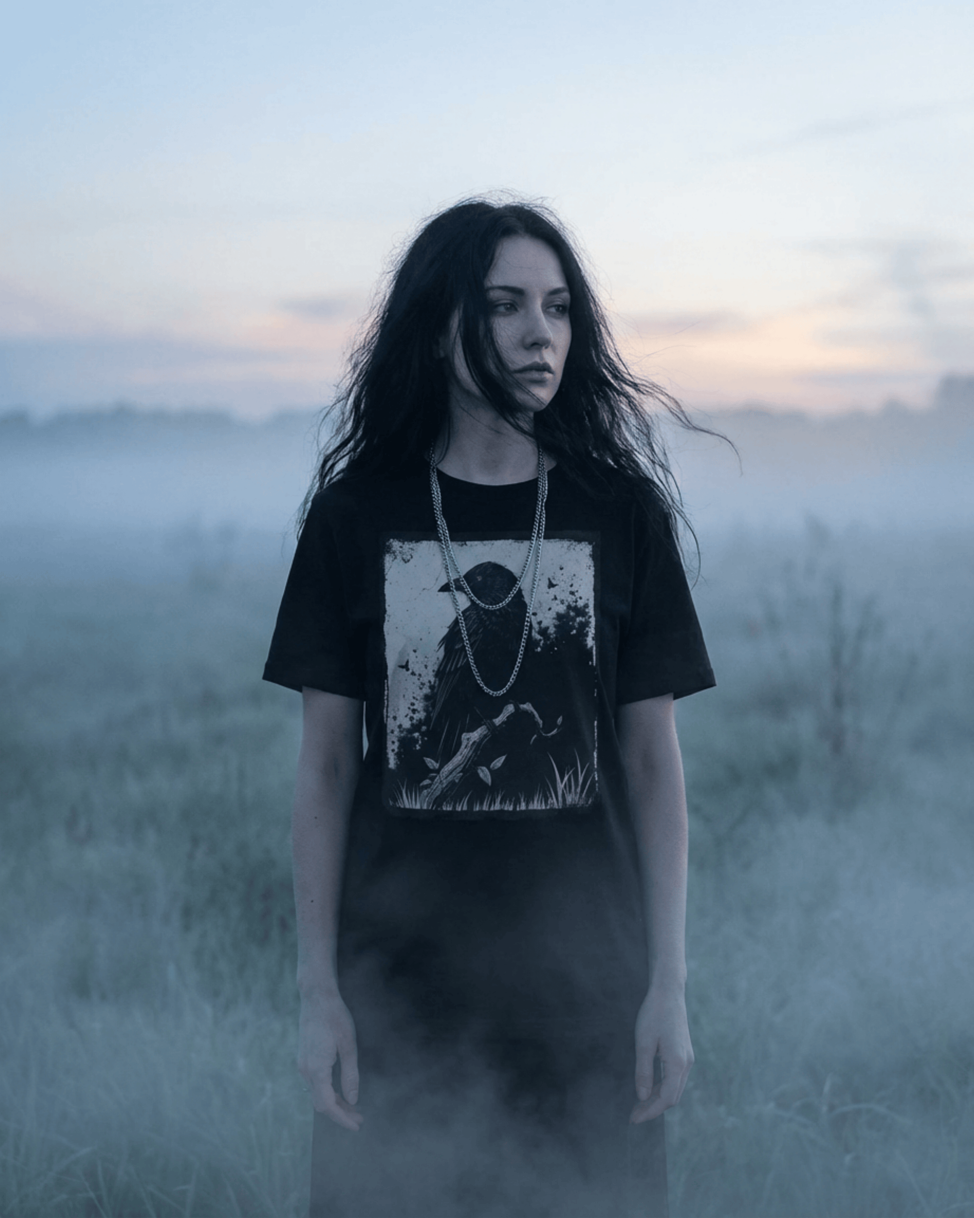A woman with dark, flowing hair models the Goth Alternative Style Shirt in a misty field at dusk, capturing a dark academia aesthetic. This grunge witchy crow t-shirt for women is styled with a silver chain.